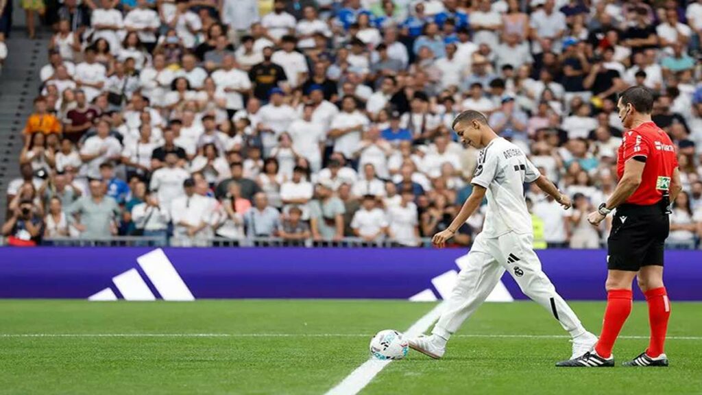 Un jeune marocain en survêtement du Real Madrid salue le public du stade Santiago Bernabéu, sous les applaudissements des joueurs et arbitres.